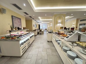 Un espace buffet avec plusieurs présentoirs de nourriture, un coin repas avec des tables et des chaises, et une vue sur la salle à manger en arrière-plan.