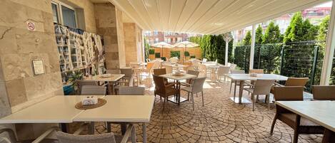 An outdoor cafe with tables and chairs, a tiled floor, and a view of buildings and trees.