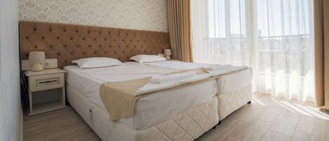 Una habitación de hotel con una cama grande, una mesita de noche, una lámpara y un ventanal con cortinas.