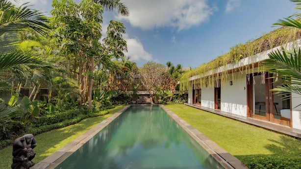 A swimming pool with a statue, surrounded by lush greenery and a building with large windows. - Villa SS Chang (Canggu)