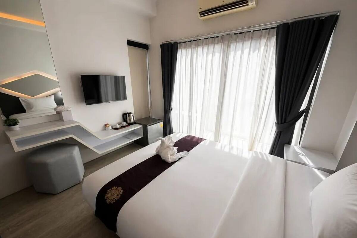 Habitación de hotel con cama, escritorio, televisor de pantalla plana, ventana con cortinas y una pequeña planta en maceta.