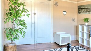 Una habitación con piso de madera, una puerta blanca, una pequeña mesa blanca, una silla negra, una planta en maceta y un adorno en la pared.