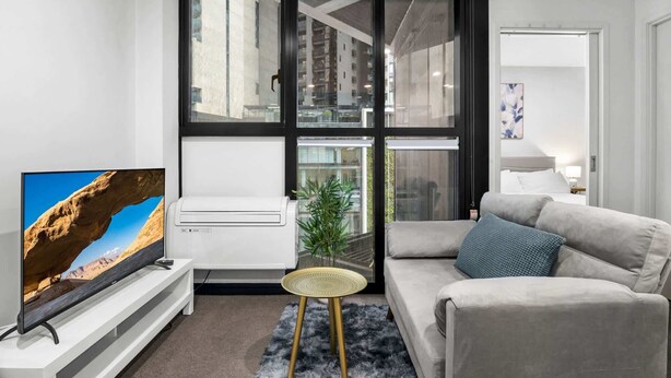 Una sala de estar moderna con un televisor de pantalla plana, un sofá, una pequeña mesa redonda y un ventanal con vistas a un edificio.