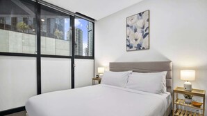 Habitación de hotel con una cama grande, ventana con vista a edificios, un cuadro en la pared y una mesita de noche con lámpara.