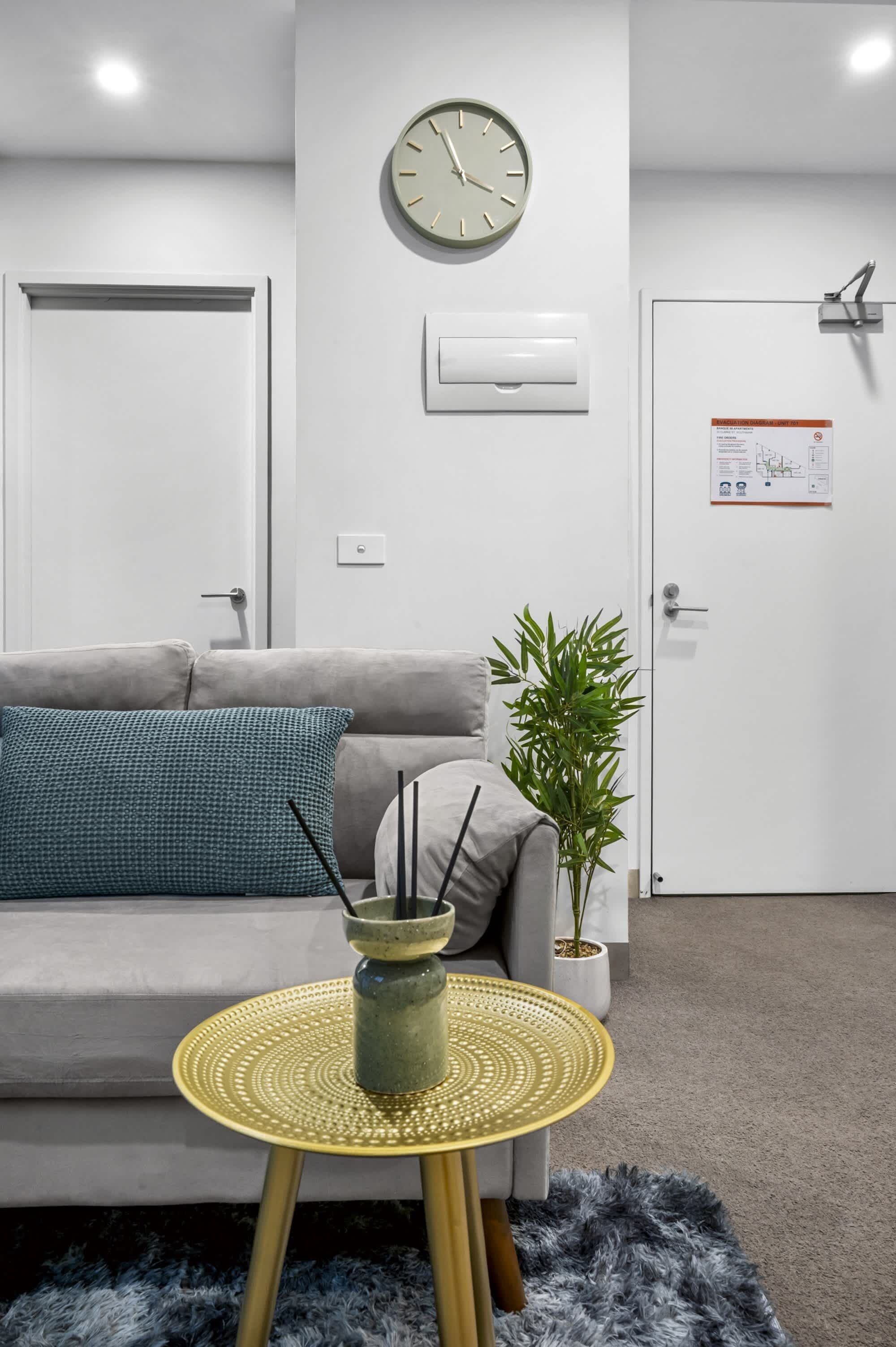 A modern living room with a sofa, a round table with a yellow base, a wall clock, a potted plant, and a door with a notice.