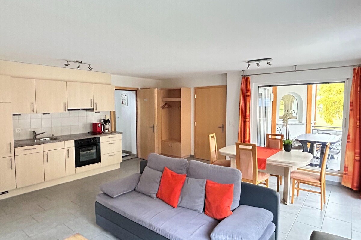 A compact living space with a kitchenette, a sofa, a dining table, and a view through a window.