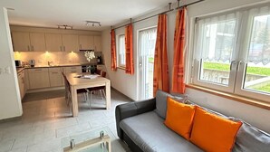 A modern kitchen with white cabinets, a dining table, and a sofa with orange cushions. - Bedroom Retreat in Saas Valley (Saas Grund)