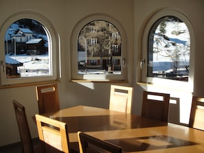 Ein Essbereich mit einem runden Tisch und vier Holzsesseln. Drei Bogenfenster bieten Blick auf eine verschneite Landschaft und Gebäude.
