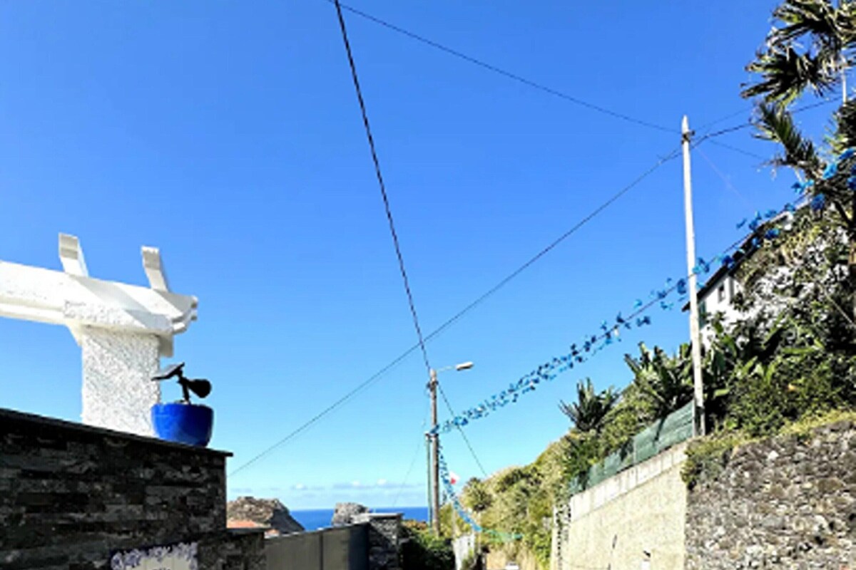 Une rue étroite bordée d’un mur de pierre, un pot bleu posé sur une structure blanche et une enseigne fixée au mur.