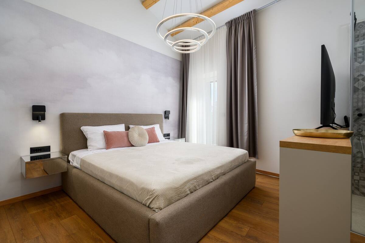 A modern bedroom with a large bed, bedside tables, a wall-mounted light fixture, and a view of the outside through a window.