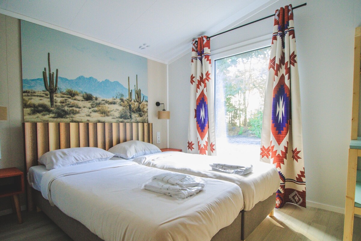 A double bed with white linens, a nightstand, a lamp, and a desert mural on the wall.