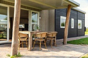 Un patio al aire libre con una mesa de madera y sillas de mimbre, una planta en maceta y una casa con ventanas amplias y fachada oscura.