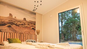 A bedroom with a large bed, a wall mural, a window with a view of trees, and a ceiling with a wooden panel design. - Modern Holiday Home With Infrared Sauna (Slagharen)
