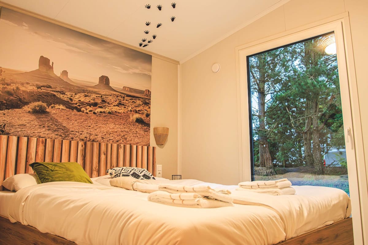 Un dormitorio con una cama grande, un mural en la pared, una ventana con vistas a árboles y un techo con diseño de paneles de madera.