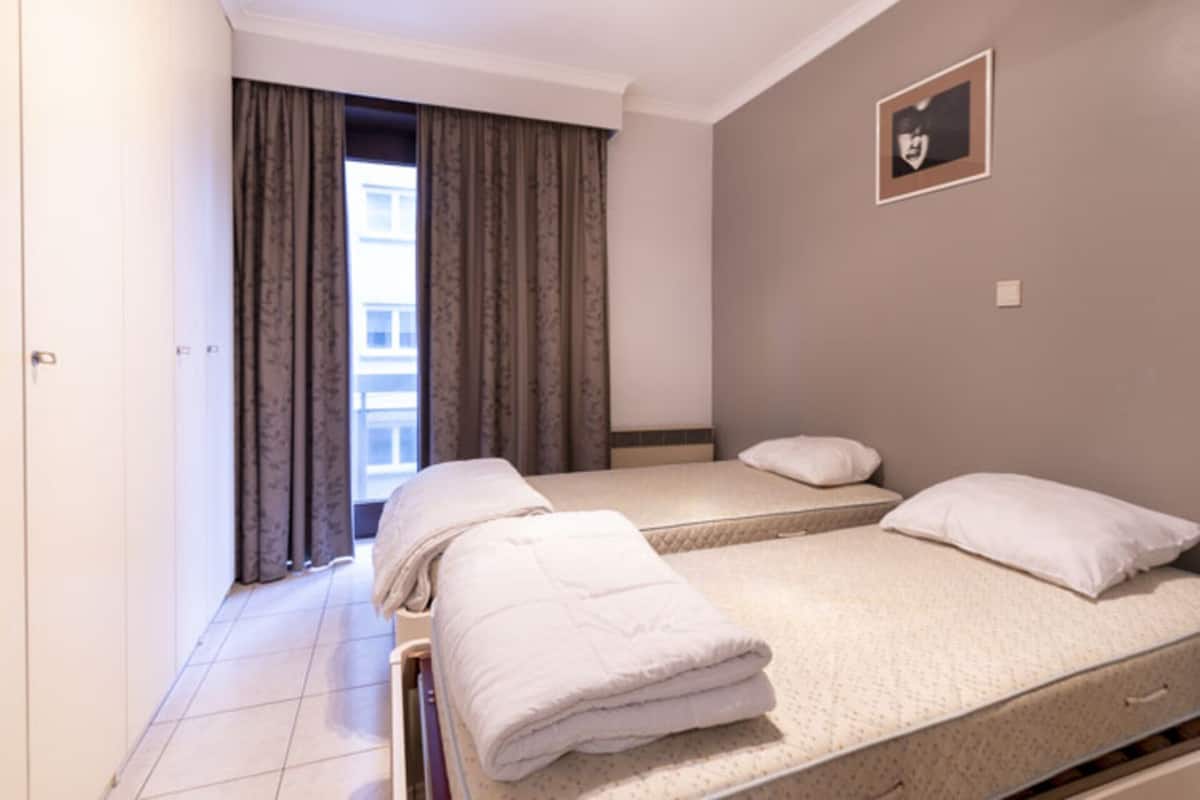Ein Hotelzimmer mit zwei Einzelbetten, einem großen Fenster mit Vorhängen, einem Bild an der Wand und einer Tür links.