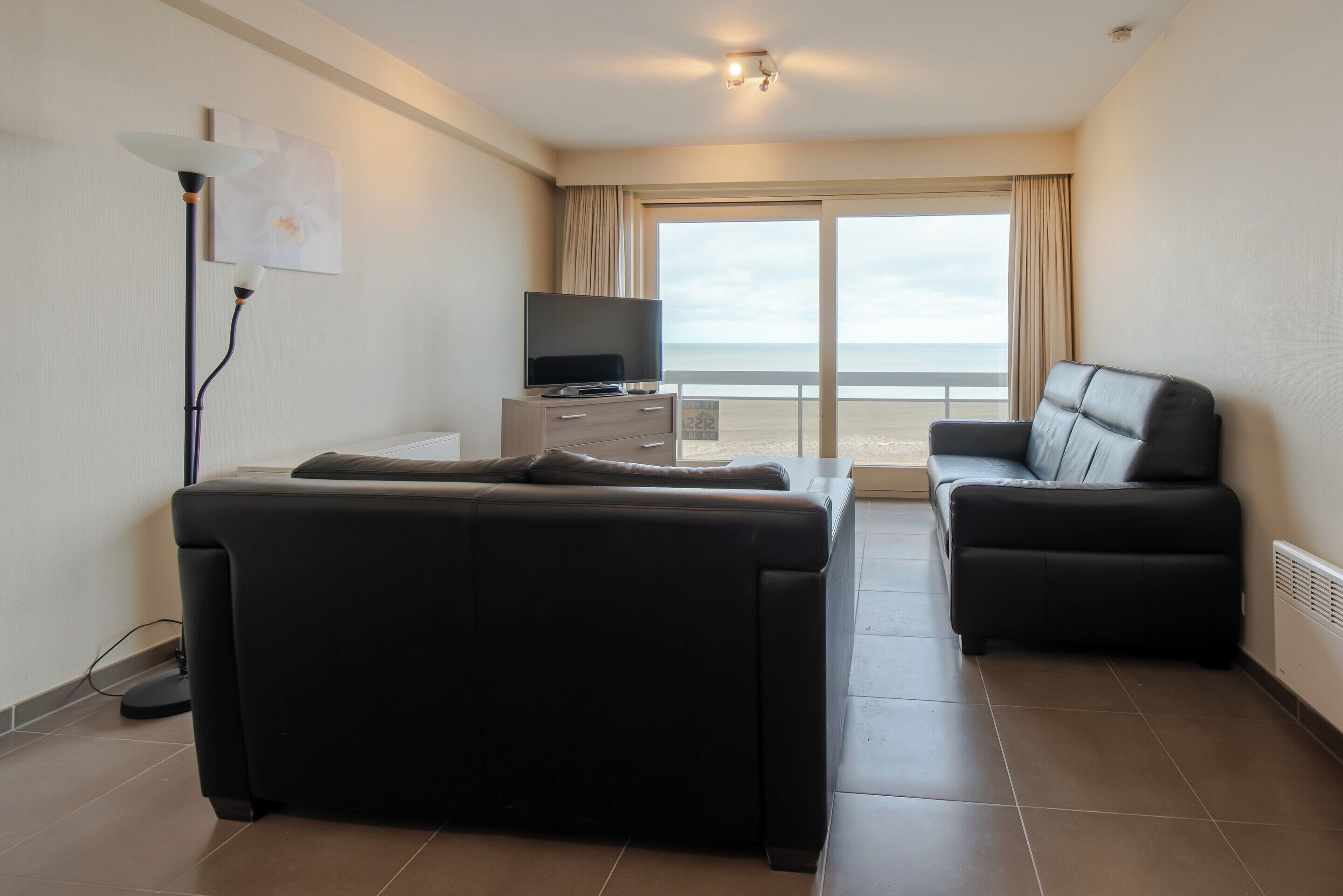 A modern hotel room with a large sofa, a smaller armchair, a flat-screen TV on a white stand, and a view of the ocean through a large window.