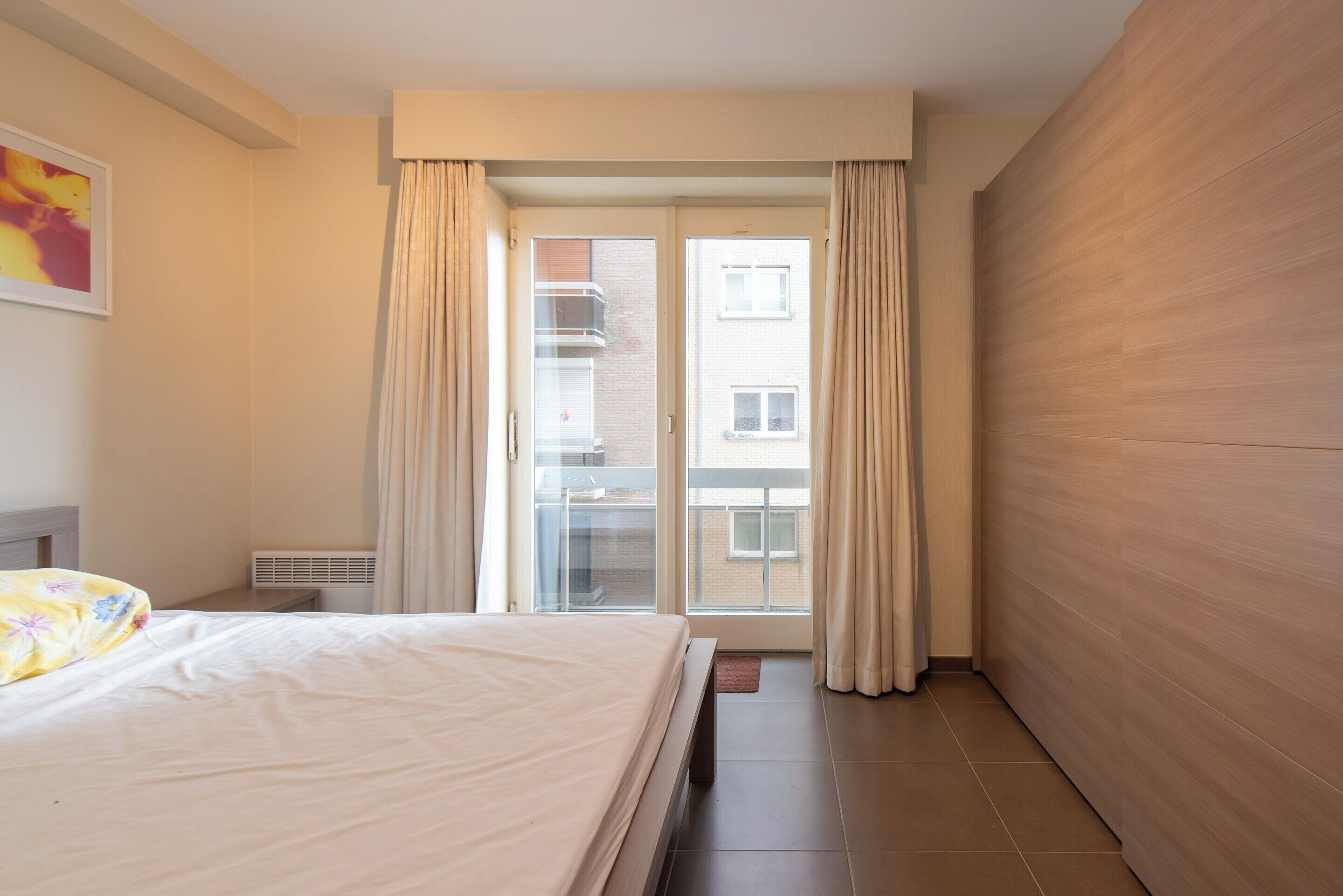 Ett hotellrum med en säng, ett fönster med gardiner, ett element och utsikt över byggnaden utanför.