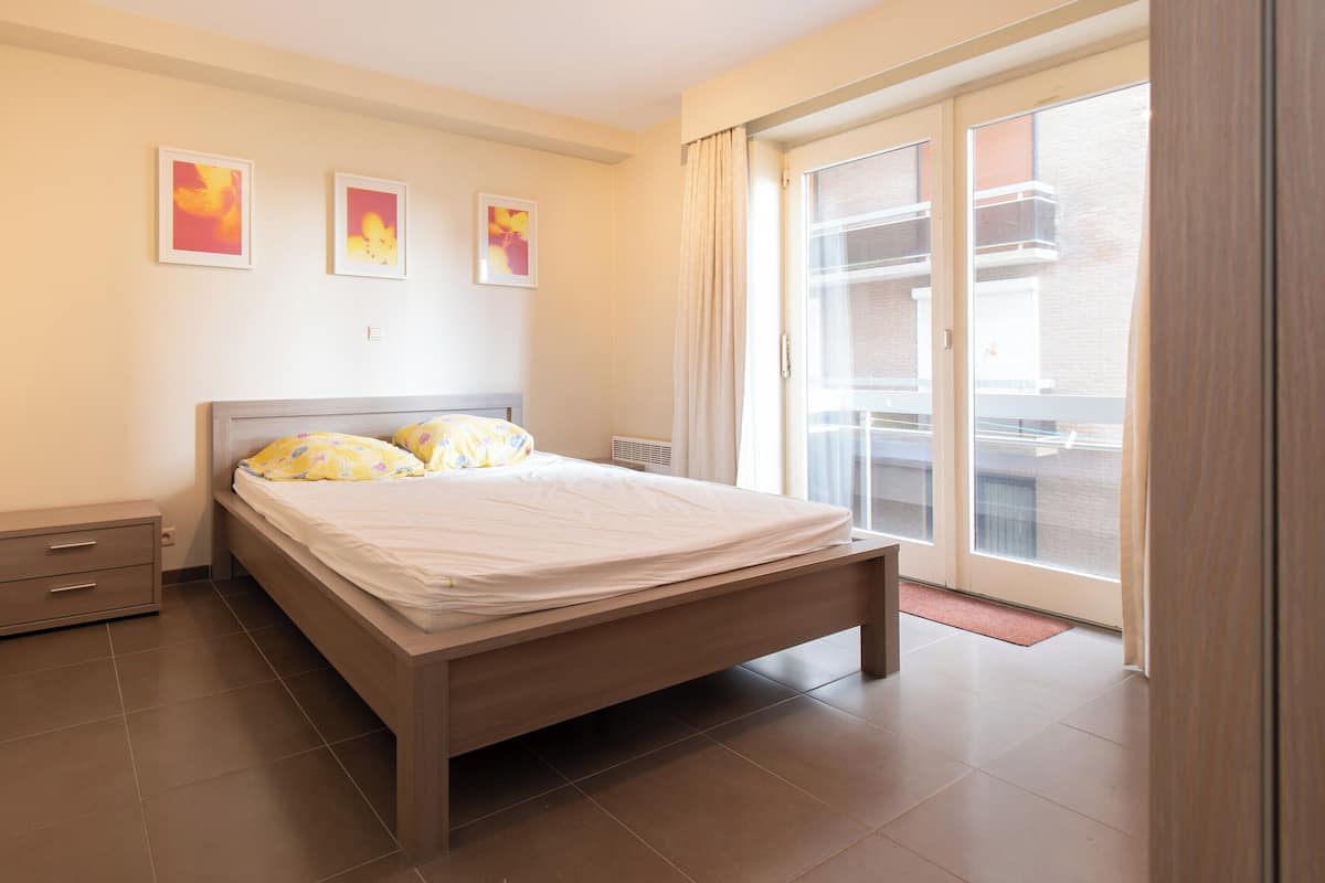 Ein Schlafzimmer mit einem Bett, einem Nachttisch, einem Fenster mit Blick auf einen Balkon und drei gerahmten Bildern an der Wand.