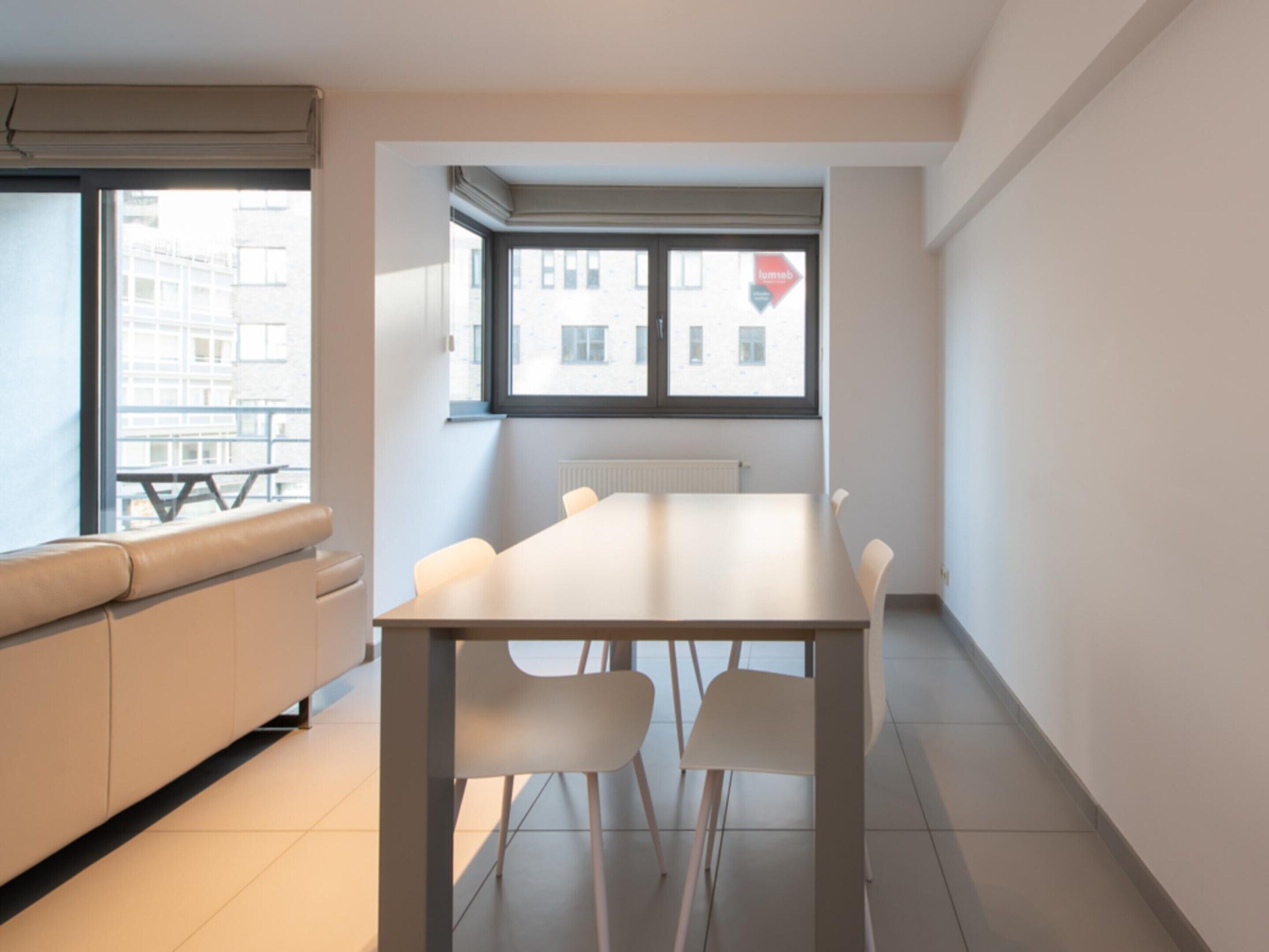 Ett modernt, minimalistiskt rum med ett vitt bord och stolar, en beige soffa och ett fönster med utsikt över byggnader.