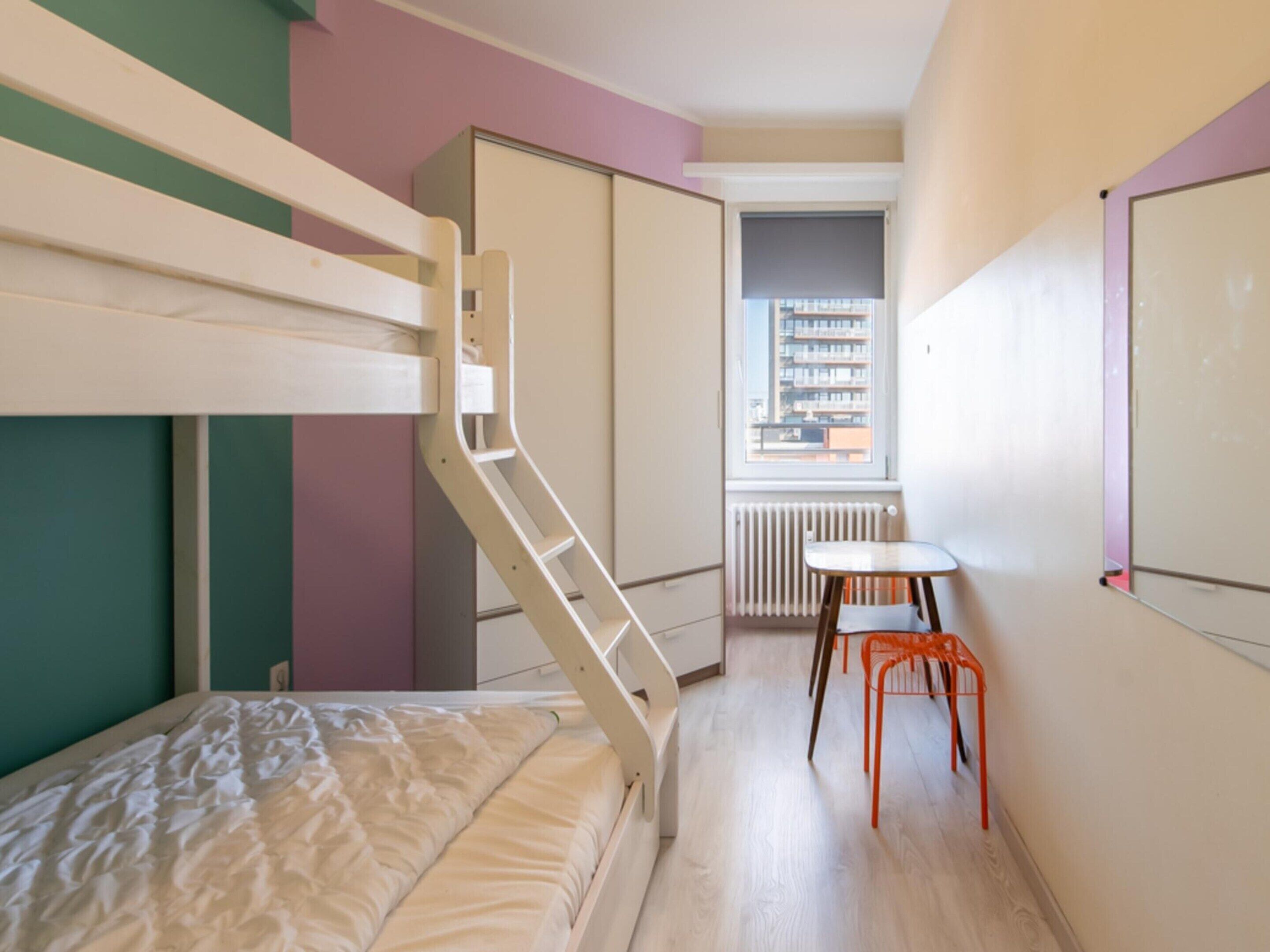 A bunk bed with a ladder, a small table, a chair, and a radiator in a room with a window and a door leading to another room.
