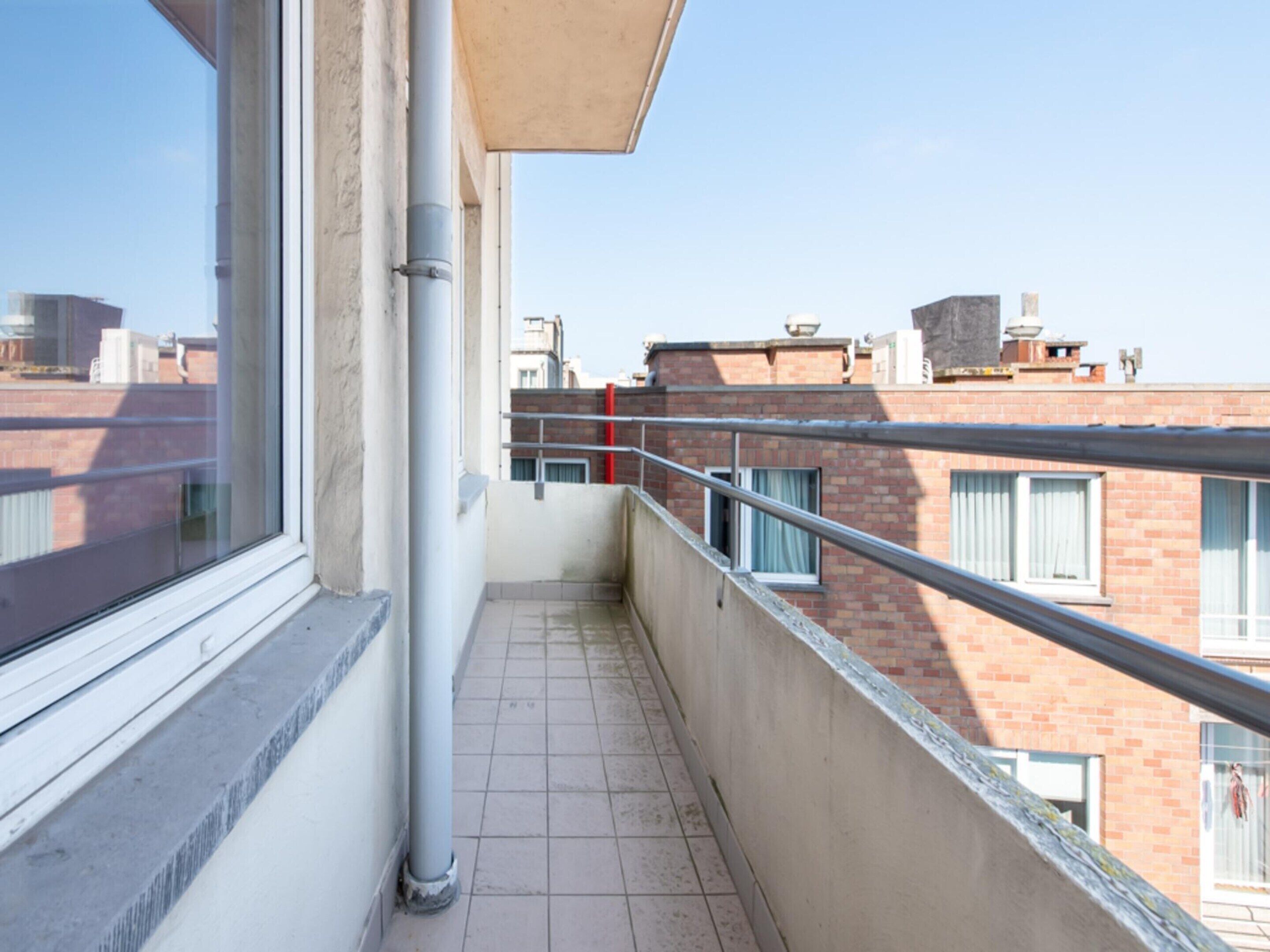 Un balcón estrecho con barandilla de metal, una ventana y vistas a edificios cercanos.