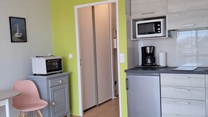 Una cocina moderna con microondas, cafetera y un refrigerador pequeño. En la sala de estar hay una mesa de vidrio y una silla rosa.