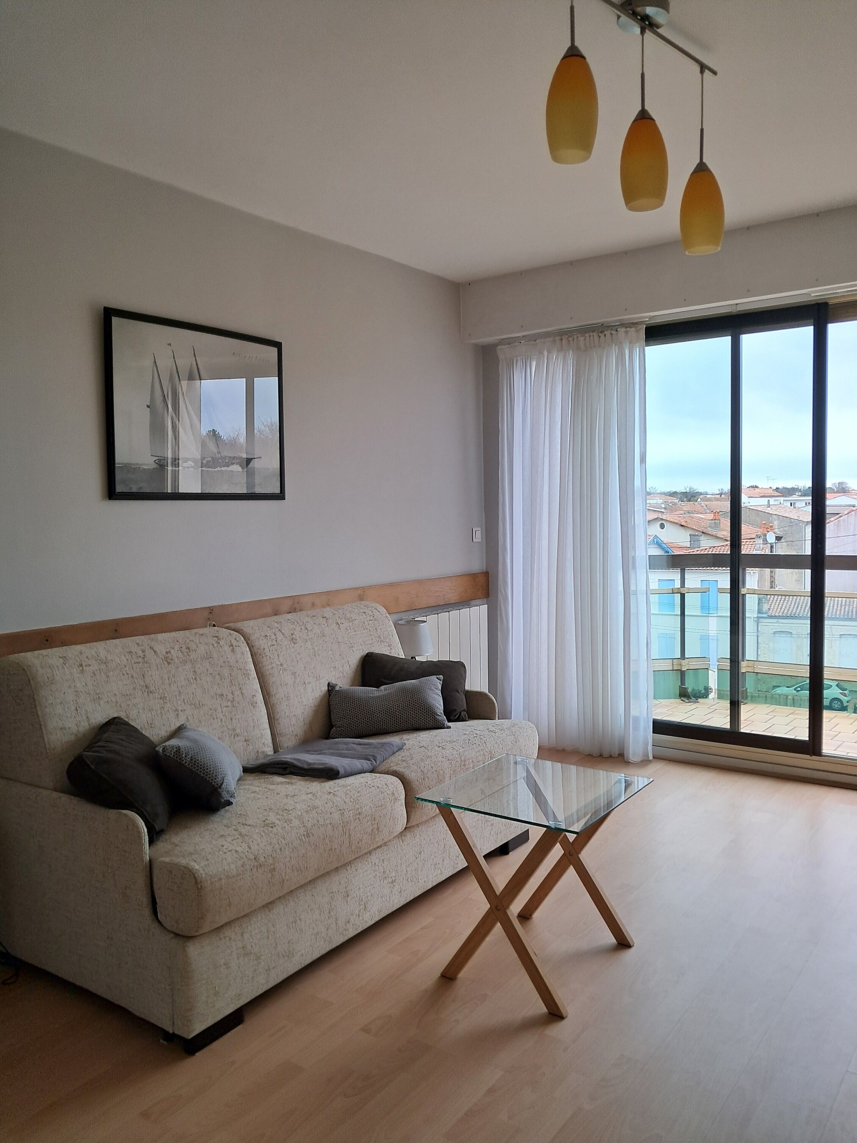 Uma sala de estar com um sofá, uma mesa de centro de vidro, um quadro na parede e uma janela grande com cortinas.