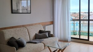 Una sala de estar con un sofá, una mesa de centro de vidrio, un cuadro en la pared y un amplio ventanal con cortinas.