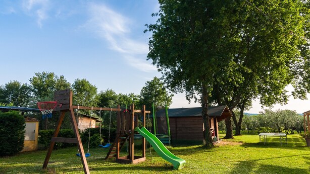Ein Spielplatz mit Rutsche und Schaukeln, umgeben von Bäumen und einem Rasen.