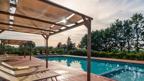 Ein Swimmingpool mit Liegestühlen und einer überdachten Liegefläche, umgeben von Bäumen.