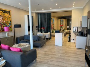 Una sala moderna con un sofá gris, una mesa de centro y un comedor con mesa y sillas. Hay una cocina con estufa y lavaplatos.