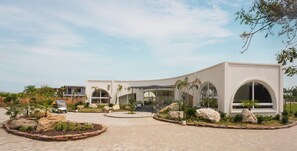 Um edifício moderno com fachada curva, grandes janelas em arco e uma entrada arborizada com pedras e plantas.