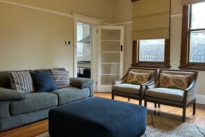 A living room with a sofa, ottoman, and two armchairs, a closet with a sliding door, and a window with blinds. - Heritage Charmer | Roomy and cosy | Central Orange (Orange)