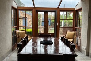 Un comedor con mesa y sillas de madera, ventanas amplias y vista a un edificio de ladrillo.