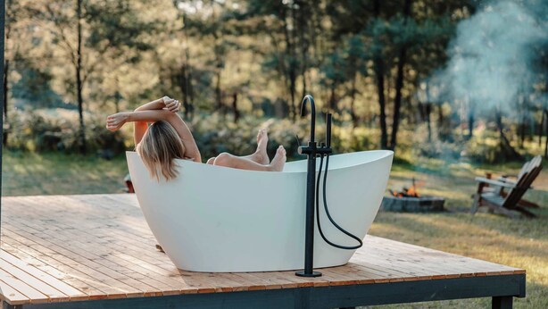Une personne se détend dans une baignoire blanche sur une terrasse en bois, avec un foyer au fond.