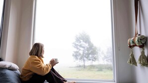 Une personne est assise près d’une fenêtre, d’où l’on aperçoit un arbre, une décoration en macramé suspendue et un coussin sur un lit.