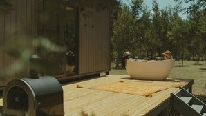 Une terrasse en bois avec un barbecue, un grand saladier blanc et un décor rustique agrémenté d’arbres.