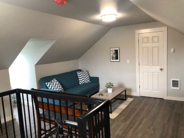 Un espacio tipo loft con techo inclinado, un sofá verde oscuro con cojines estampados, una mesita blanca y una puerta blanca.