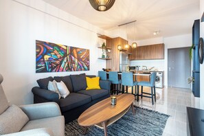 A modern living room with a sofa, coffee table, and chairs, a kitchen with wooden cabinets and a dining area, and a colorful abstract painting on the wall. - Sunshine 1-BR Apt. & Blacony in Larnaca (Larnaca)