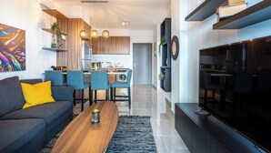 A modern living room with a sofa, coffee table, and TV. There is a kitchen in the background with a dining area. - Sunshine 1-BR Apt. & Blacony in Larnaca (Larnaca)