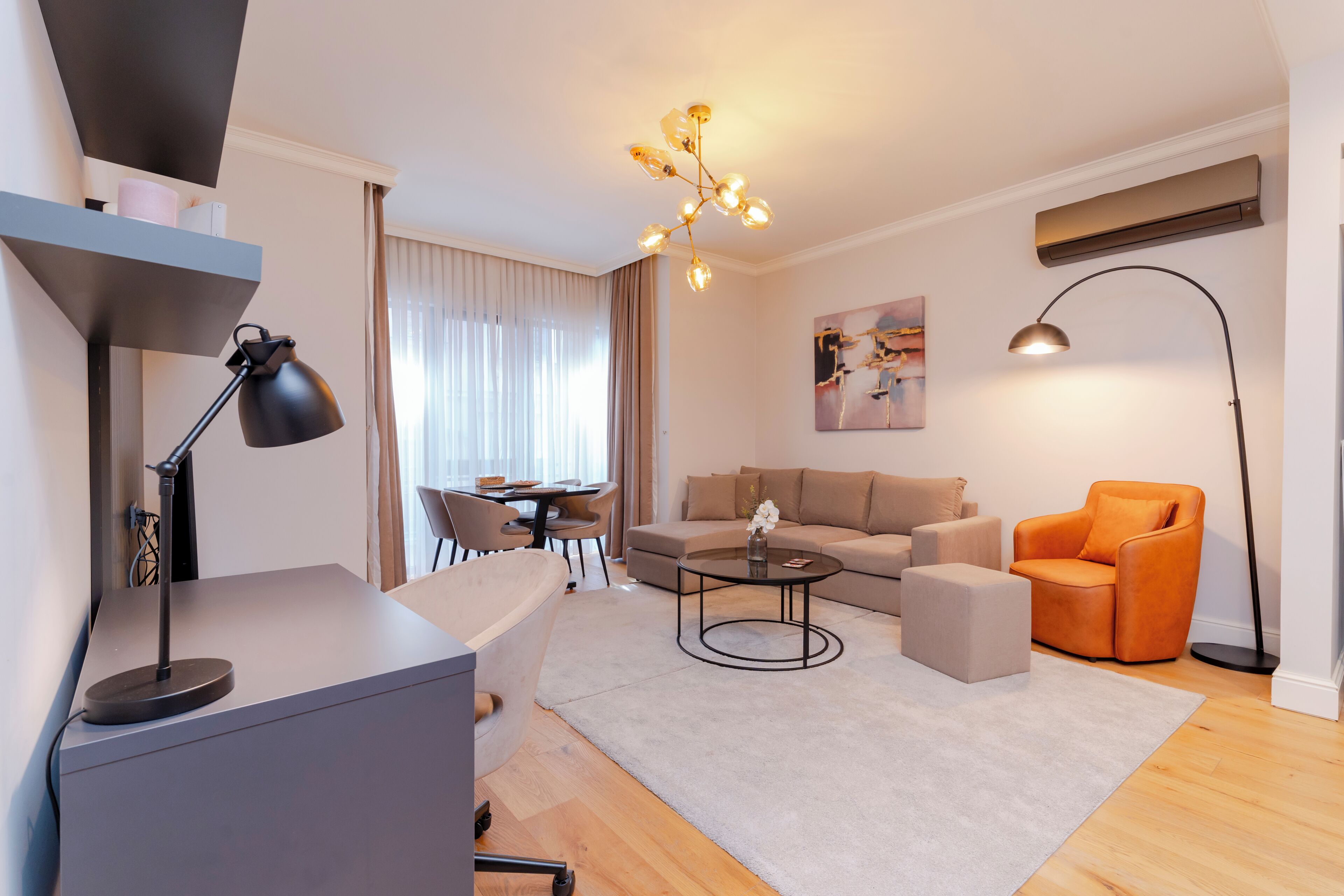 Ein modernes Wohnzimmer mit einer Couch, einem Couchtisch und einem orangefarbenen Sessel. Es gibt einen Schreibtisch mit einer Lampe, einen Essbereich und ein Bild an der Wand.