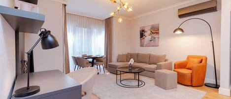 Ein modernes Wohnzimmer mit einer Couch, einem Couchtisch und einem orangefarbenen Sessel. Es gibt einen Schreibtisch mit einer Lampe, einen Essbereich und ein Bild an der Wand.