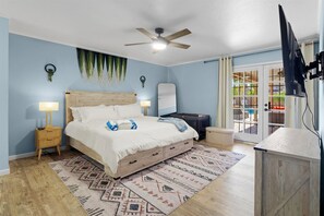 Ein Schlafzimmer mit Bett, Nachttischlampen, Deckenventilator, großem Spiegel, Fernseher und Blick ins Freie durch eine Schiebetür.