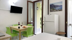 A hotel room with a bed, a dining table with chairs, a TV mounted on the wall, a refrigerator, and a painting of a landscape. - Porto Letizia Mascagni 1, Porlezza, Italy (Porlezza)