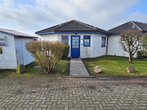 A white building with a blue door and windows, a paved driveway, and a small garden with a tree and shrubs.