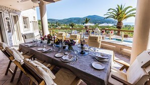 A dining area with a table set for four, outdoor chairs, a ceiling fan, and a view of mountains and greenery. - Epidavros Villas, Elia Garden Retreat, Shared Pool (Nea Epidavros)