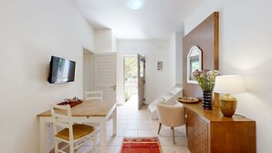 Un comedor con mesa de madera, sillas blancas, televisor montado en la pared y un florero con flores sobre un mueble de madera.