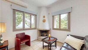A living room with a fireplace, a red chair, a sofa, and large windows. - Epidavros Villas -  Dafni Stone & Garden Retreat (Nea Epidavros)