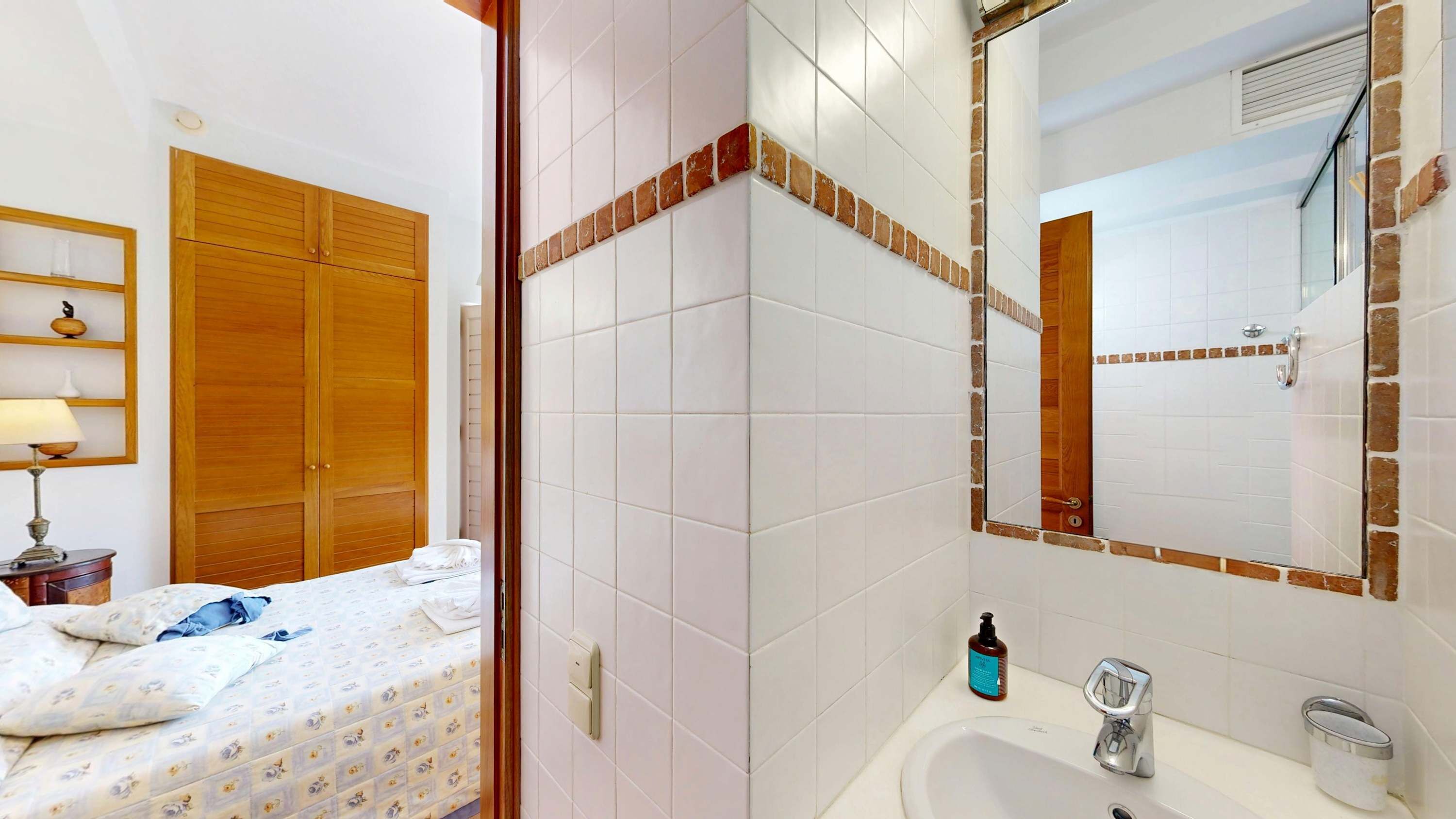 Une chambre d’hôtel comprenant un lit, une armoire en bois, une salle de bain avec un lavabo et un miroir, et une étagère avec des articles de toilette.