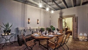 Un coin repas avec une table en bois dressée pour six personnes, entourée de six chaises. Deux appliques murales sont fixées au-dessus de la table, et un banc avec des coussins se trouve à gauche.